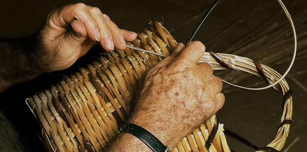 Primer plano de las manos de una persona tejiendo una cesta con finas tiras de material natural de color claro. La persona lleva una muñequera oscura en la muñeca izquierda.