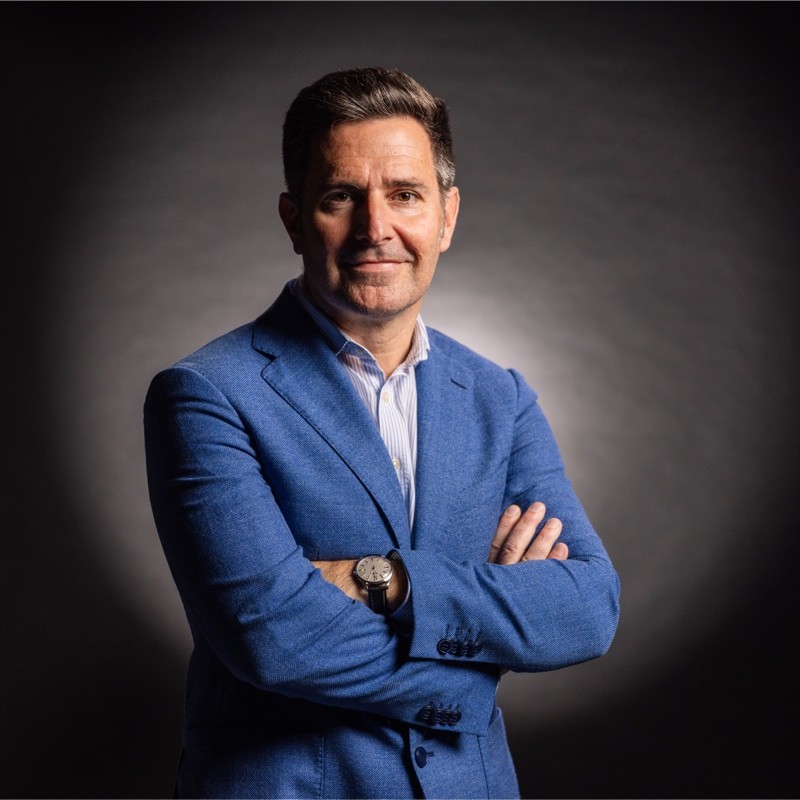 Un hombre con traje de chaqueta azul y camisa de rayas sonríe confiado con los brazos cruzados sobre un fondo oscuro y suavemente iluminado.