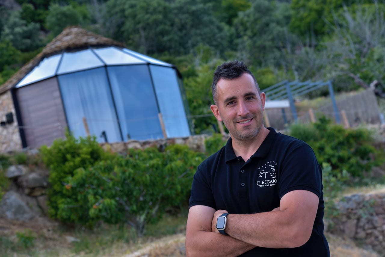 Un hombre con el pelo corto y oscuro y un polo negro sonríe con los brazos cruzados delante de un moderno edificio en forma de cúpula rodeado de vegetación.