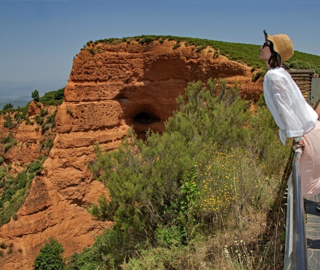 Una mujer con sombrero de paja y gafas de sol se apoya en una barandilla, mirando unos grandes acantilados de color rojo anaranjado con vegetación verde y cielo azul al fondo. Flores silvestres y arbustos bordean el acantilado.