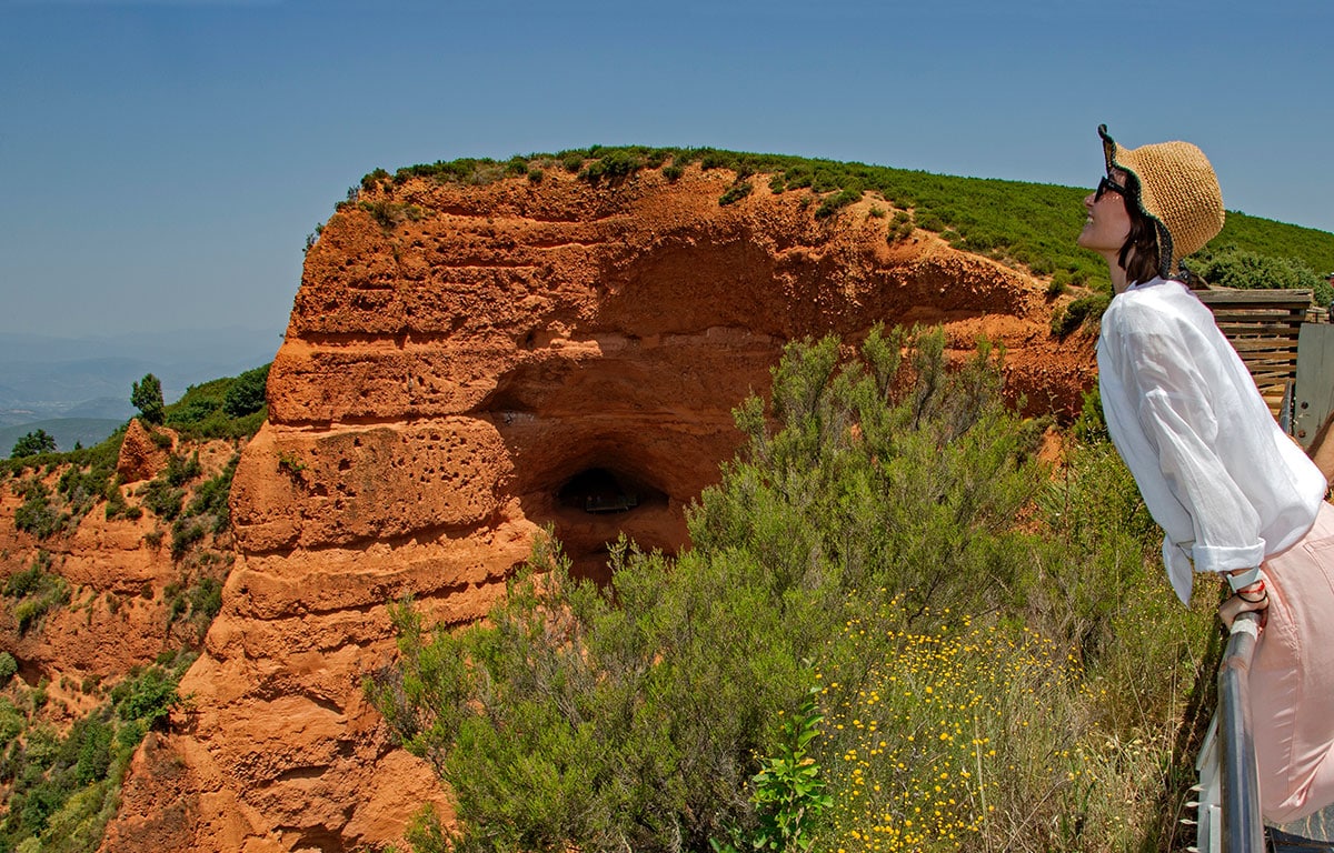 Una mujer con sombrero de paja y gafas de sol se apoya en una barandilla, admirando acantilados y cuevas de arcilla roja en Las Médulas, en España, con vegetación verde y un cielo azul despejado al fondo.