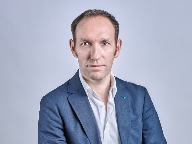 Un hombre de pelo corto castaño claro, vestido con traje de chaqueta azul y camisa de vestir blanca, posa sobre un fondo liso gris claro, mirando directamente a la cámara con expresión neutra.