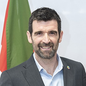 Un hombre de pelo oscuro corto y barba, vestido con traje de chaqueta y camisa azul claro, sonríe a la cámara. Al fondo se ve una bandera verde y roja.
