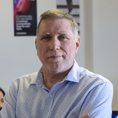 Un hombre de mediana edad, con pelo corto y canoso y camisa azul claro abotonada, está de pie en el interior de una casa con los brazos cruzados, mirando con confianza a la cámara. En el fondo se ven carteles borrosos y personas.