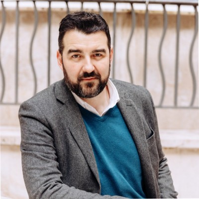 Un hombre de pelo oscuro y barba lleva una americana gris sobre un jersey azul y una camisa blanca. Está sentado frente a una pared de color claro con barandillas metálicas decorativas al fondo.