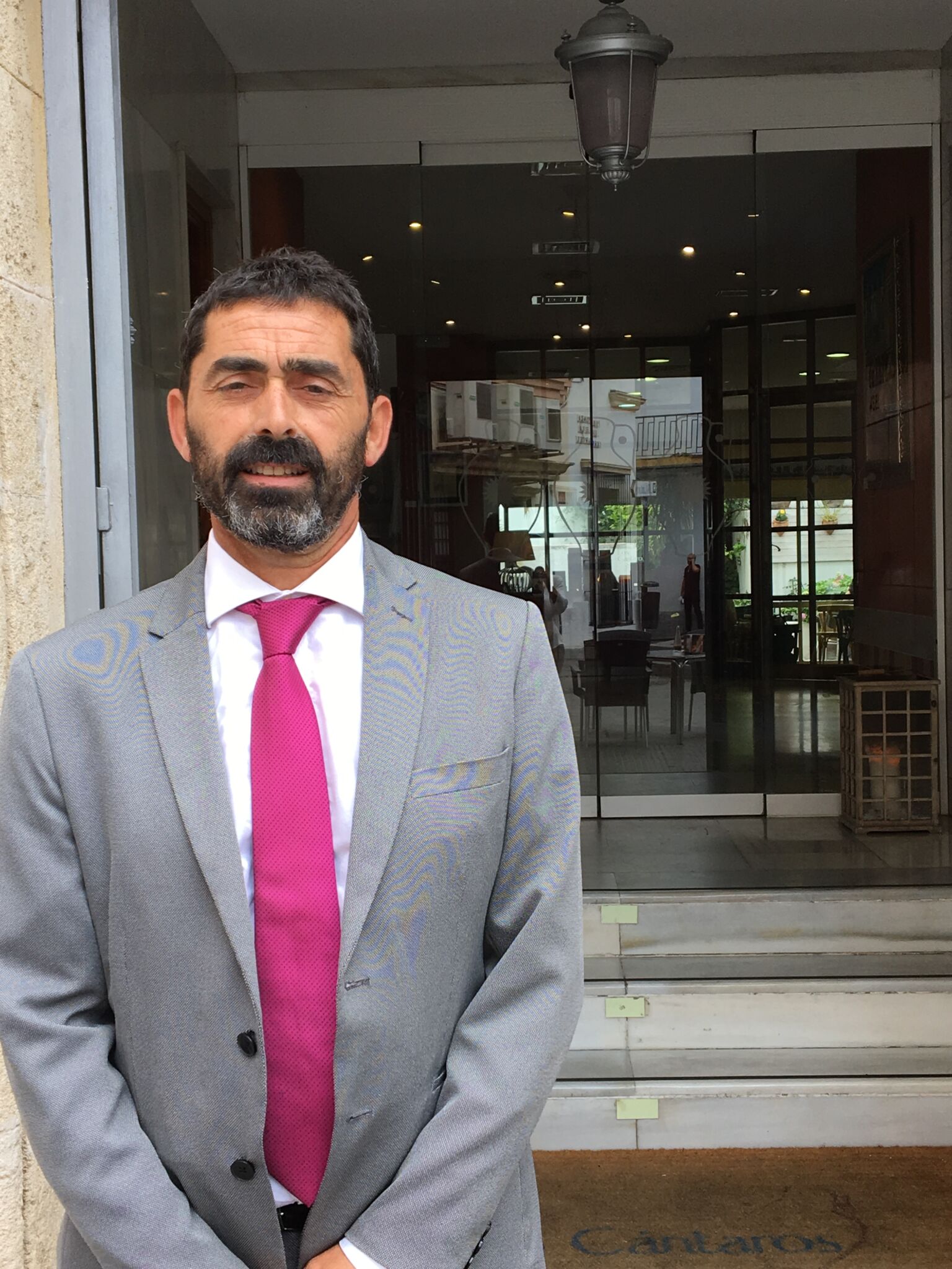 Un hombre de pelo corto y oscuro y barba, vestido con traje gris, camisa blanca y corbata rosa brillante, se encuentra frente a las puertas de cristal de la entrada de un edificio.