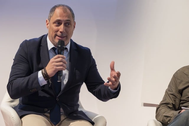 Un hombre con americana oscura y corbata se sienta en una silla blanca, habla por un micrófono y gesticula con una mano durante un acto o una mesa redonda. A su lado se ve parcialmente a otra persona.