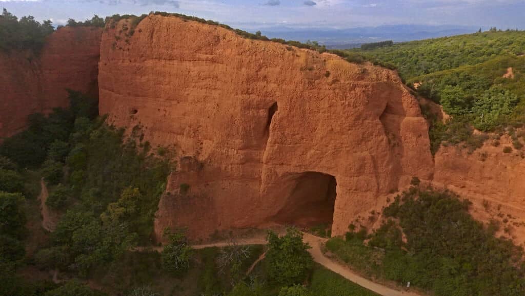 Gran acantilado rojizo con una prominente abertura en forma de cueva en su base, rodeado de vegetación y árboles, con un camino de tierra que conduce a la entrada y colinas visibles en el fondo distante.