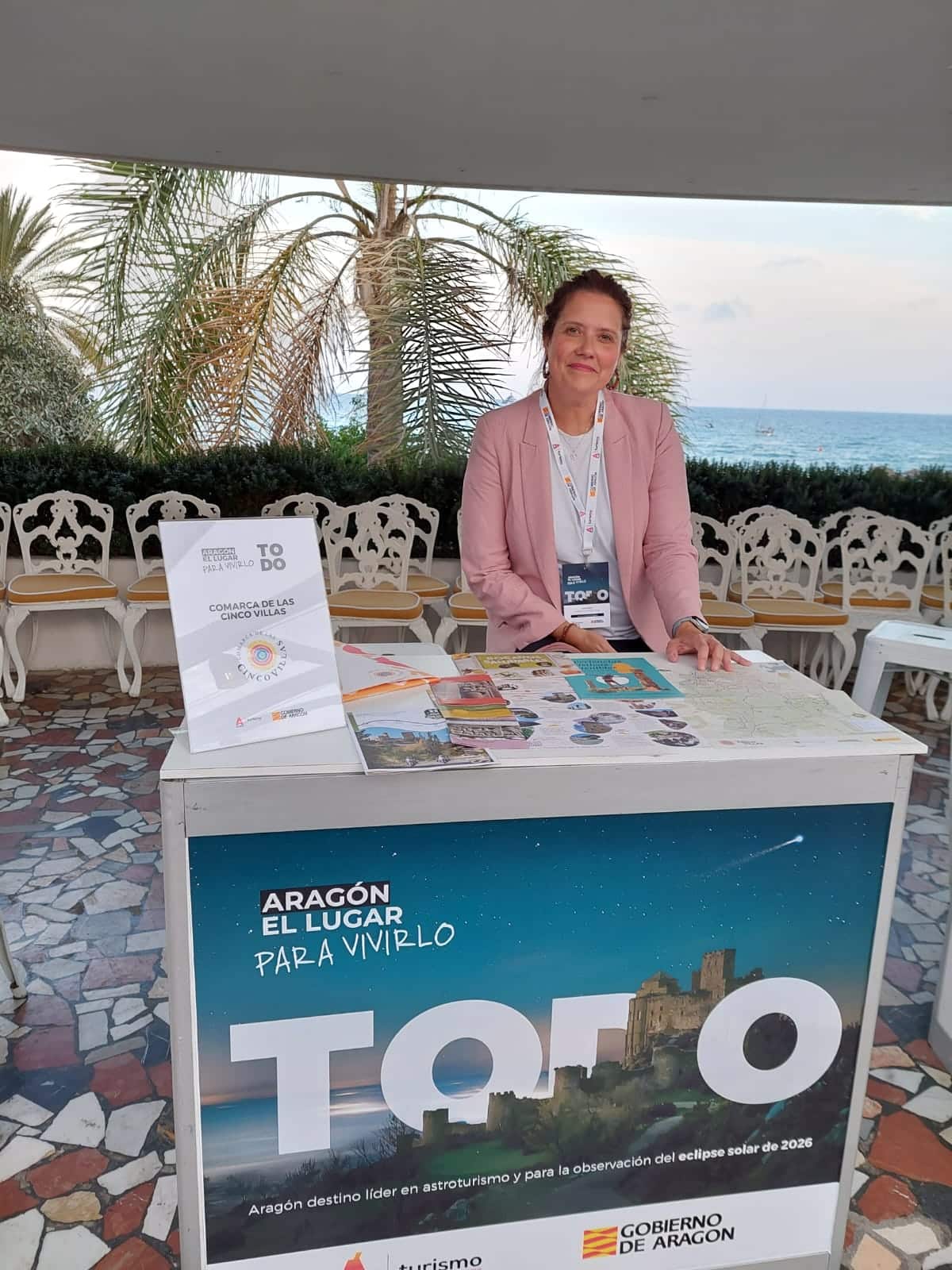 Una mujer de pie detrás de un puesto de información turística con folletos y carteles sobre Aragón, promocionando la observación del eclipse solar para 2026. Al fondo se ven palmeras y el mar.