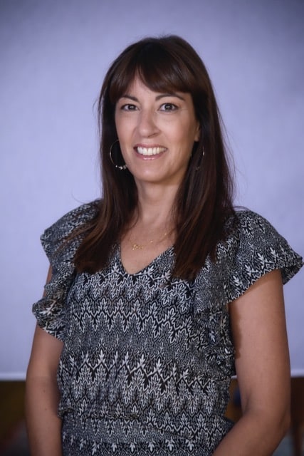 Una mujer de pelo largo y castaño con un vestido estampado de manga corta sonríe a la cámara. Está de pie delante de un fondo liso y suavemente iluminado.