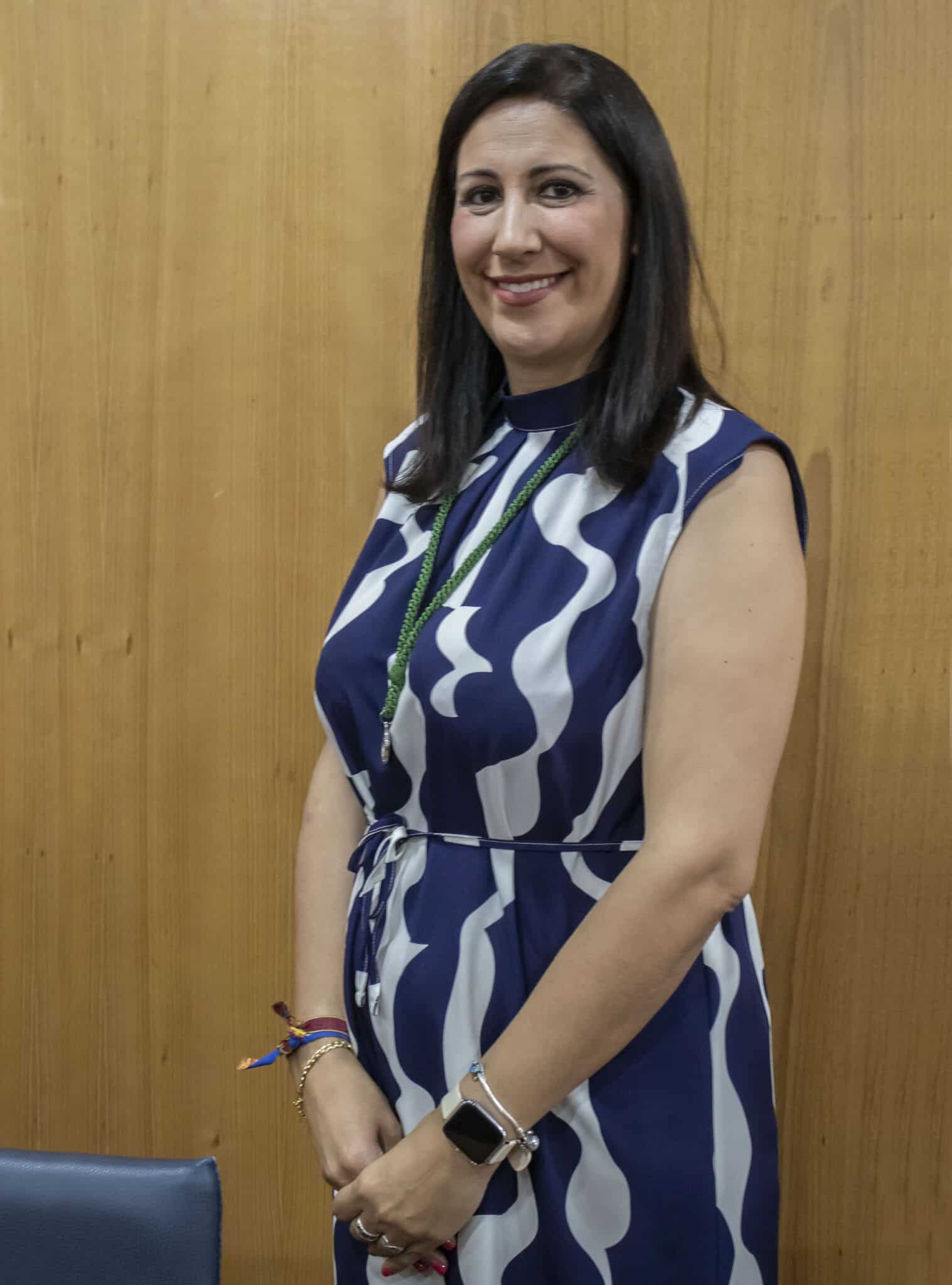 Una mujer de pelo lacio y oscuro, con un vestido estampado azul y blanco, sonríe frente a una pared de madera. Lleva pulseras, un reloj inteligente en la muñeca izquierda y un largo collar verde.