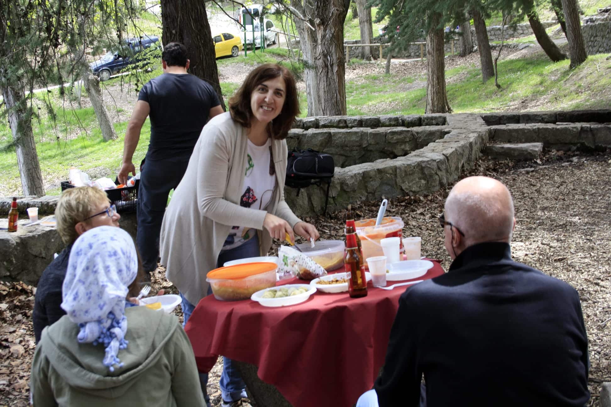 Una mujer sonríe mientras sirve comida en una mesa de picnic con otras tres personas en un parque arbolado. La mesa está cubierta con un mantel rojo, platos y bebidas. Al fondo se ven árboles y coches aparcados.