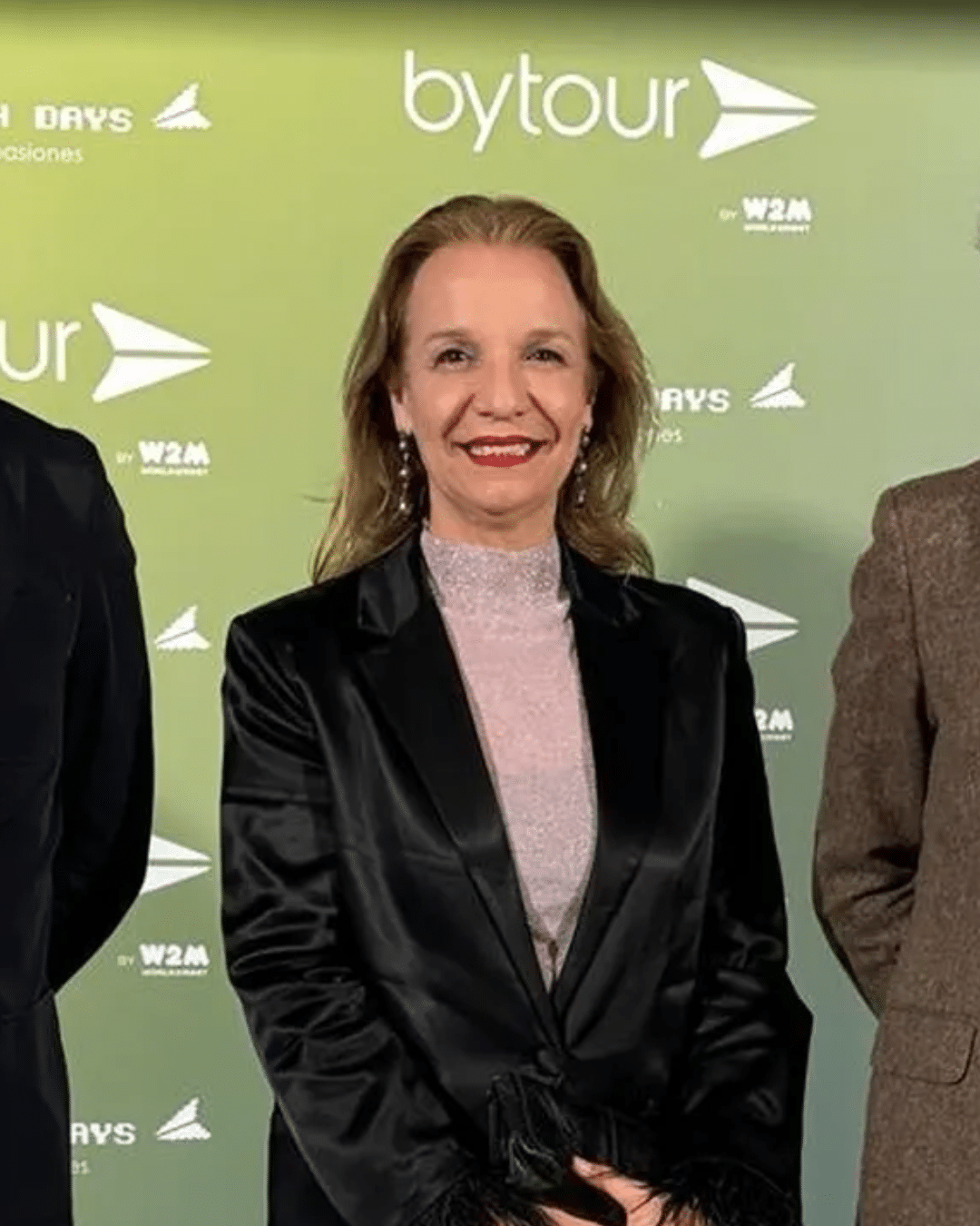 Una mujer de pelo largo y rubio, vestida con una americana negra sobre un top rosa claro, sonríe delante de un fondo verde con logotipos de bytour y otros logotipos blancos.