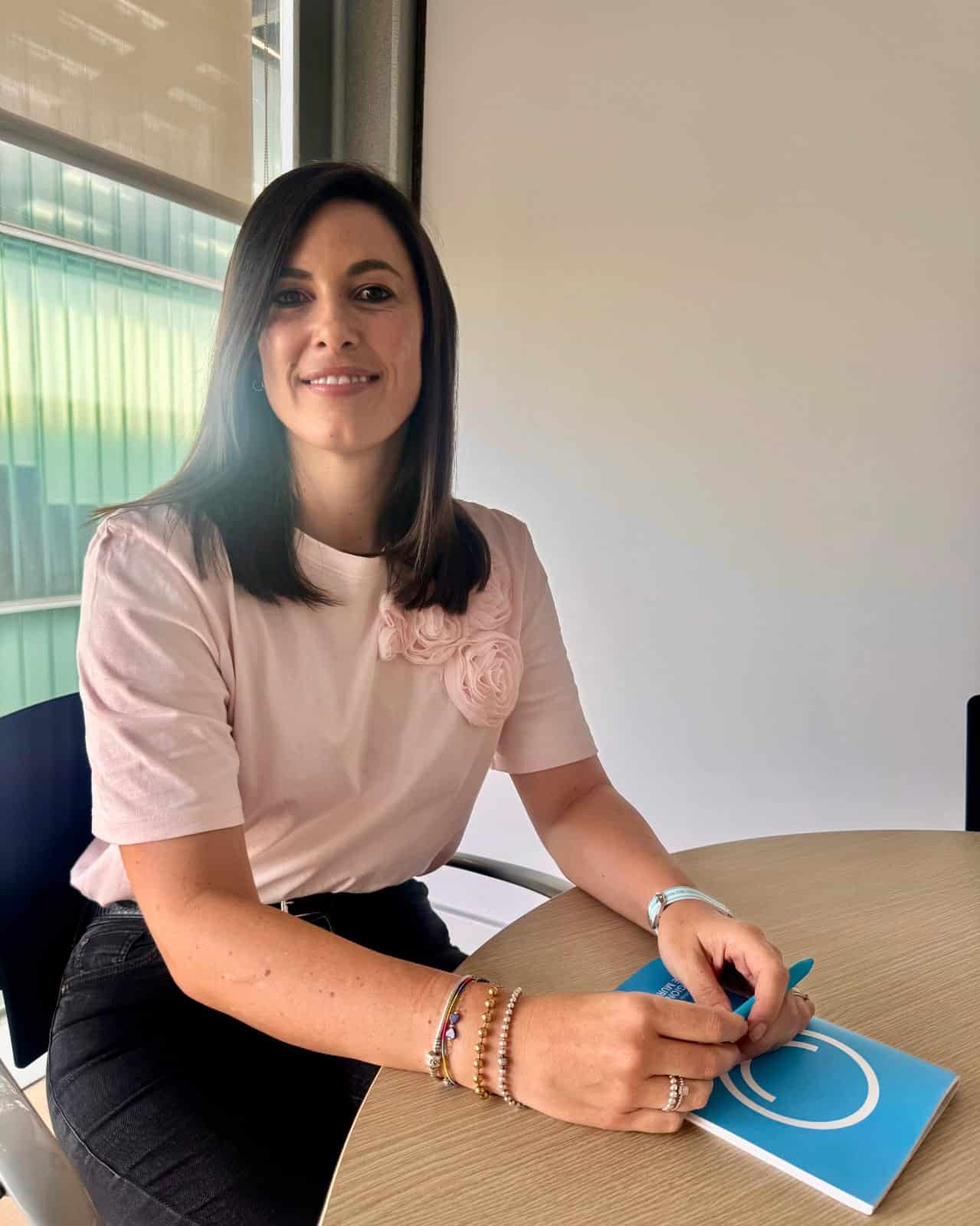 Una mujer de pelo castaño liso está sentada en una mesa redonda, sosteniendo un bolígrafo sobre un cuaderno azul. Lleva una camisa rosa claro con detalle de flor de tela y brazaletes, sonríe a la cámara en una luminosa habitación moderna.