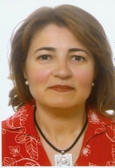 Una mujer con el pelo castaño hasta los hombros, vestida con una blusa roja con un estampado floral blanco, un collar negro con un colgante rectangular y pendientes de perlas, sonríe ligeramente delante de un fondo blanco liso.