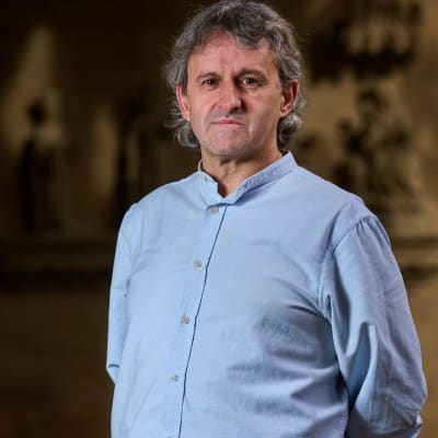 Un hombre de mediana edad con el pelo gris ondulado lleva una camisa azul claro abotonada y está de pie en un interior sobre un fondo borroso de tonos oscuros con formas indistintas. Mira directamente a la cámara con expresión neutra.