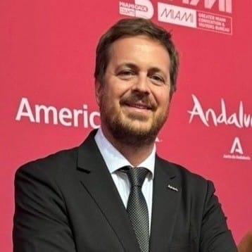 Un hombre con traje y corbata negros sonríe delante de un fondo rojo con texto y logotipos blancos, entre ellos Andalucía, Miami y América.