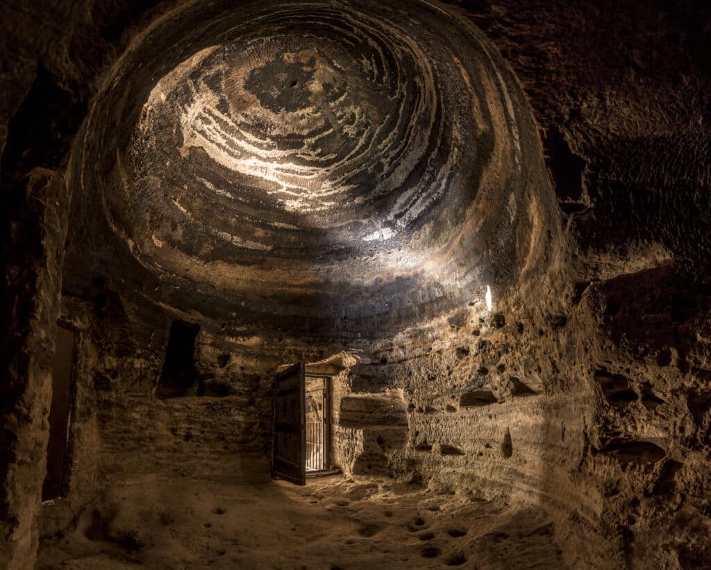 Una cueva redonda y abovedada con paredes y techo de roca texturizada y estratificada. La luz brilla a través de una puerta de madera parcialmente abierta, iluminando el áspero suelo de piedra y los cálidos tonos terrosos del interior de la caverna.