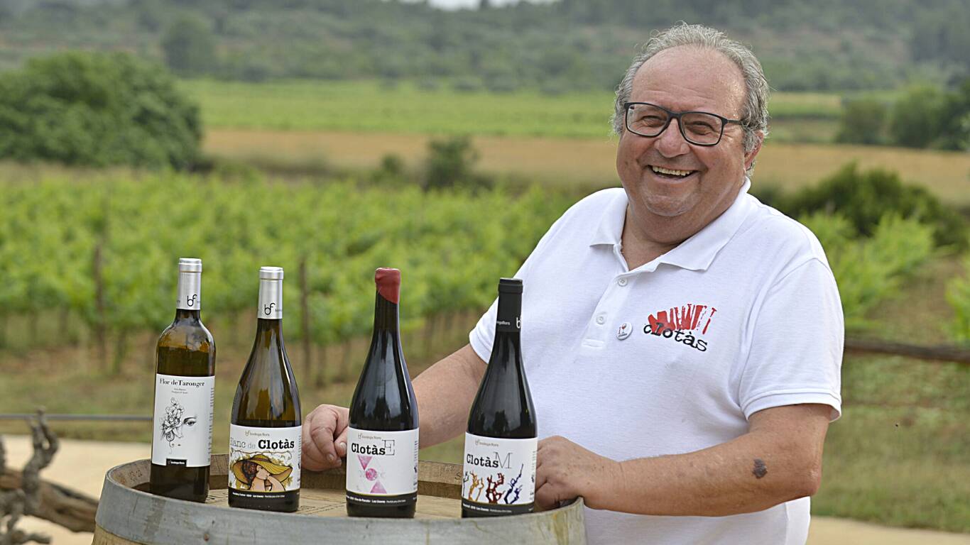 Un hombre sonriente vestido con un polo blanco se encuentra al aire libre junto a un viñedo, apoyando el brazo en un barril que muestra cuatro botellas de vino con diferentes etiquetas. Al fondo se ven exuberantes viñas y campos verdes.