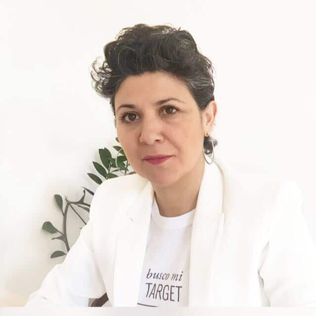 Una mujer de pelo corto y oscuro, vestida con una americana blanca y una camisa blanca con texto parcialmente visible, está sentada delante de un fondo blanco liso con una planta frondosa a su lado.