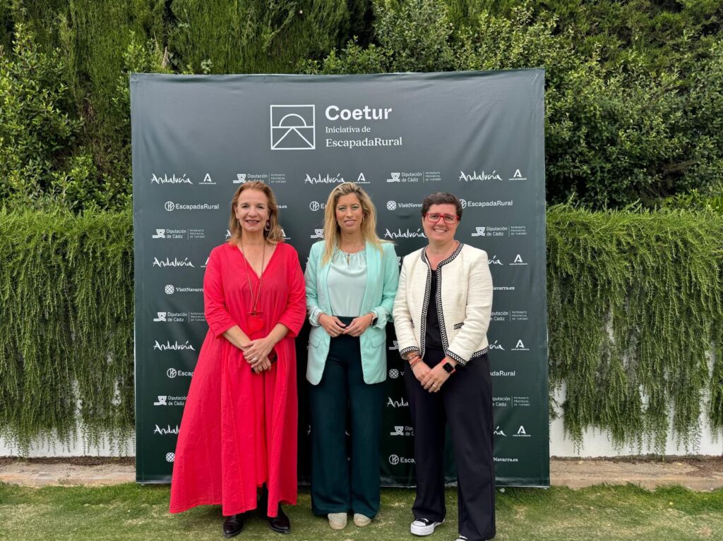 Tres mujeres sonríen, una al lado de la otra, delante de un fondo verde con el logotipo de Coetur y varios logotipos de patrocinadores. Están al aire libre, con hierba bajo los pies y vegetación detrás del telón de fondo.