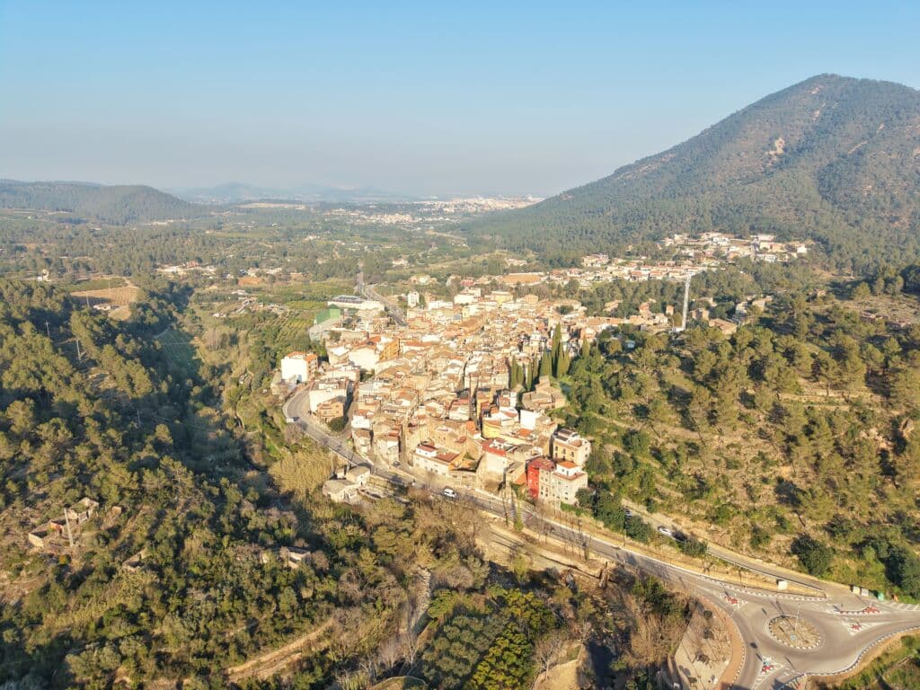 Vista aérea de una pequeña ciudad rodeada de verdes colinas y densos bosques, con una rotonda y carreteras sinuosas en primer plano y una montaña al fondo bajo un cielo despejado.