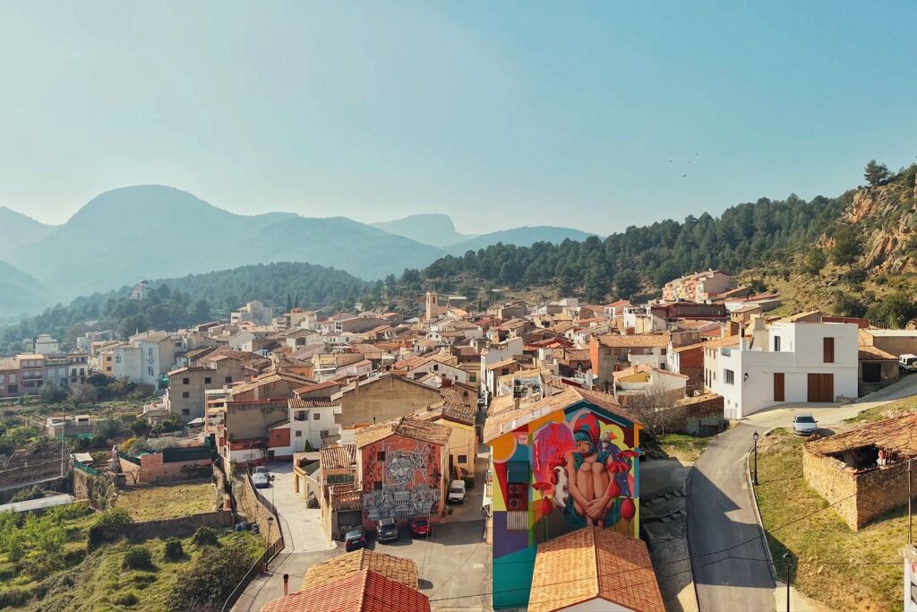 Vista aérea de una pequeña ciudad con tejados de tejas rojas rodeada de colinas y bosques; un colorido mural decora un edificio en primer plano bajo un cielo azul despejado.