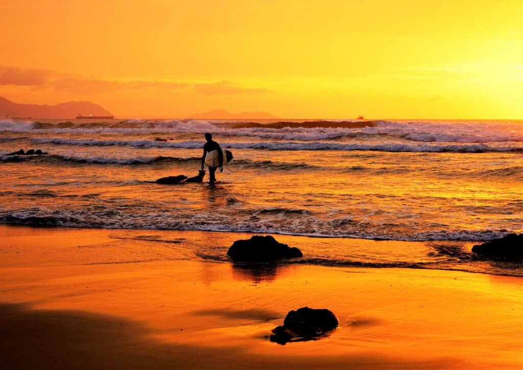 Un surfista sujeta una tabla de surf al borde del océano durante una puesta de sol de un naranja vibrante, con olas que se acercan y rocas esparcidas por la orilla de arena húmeda.