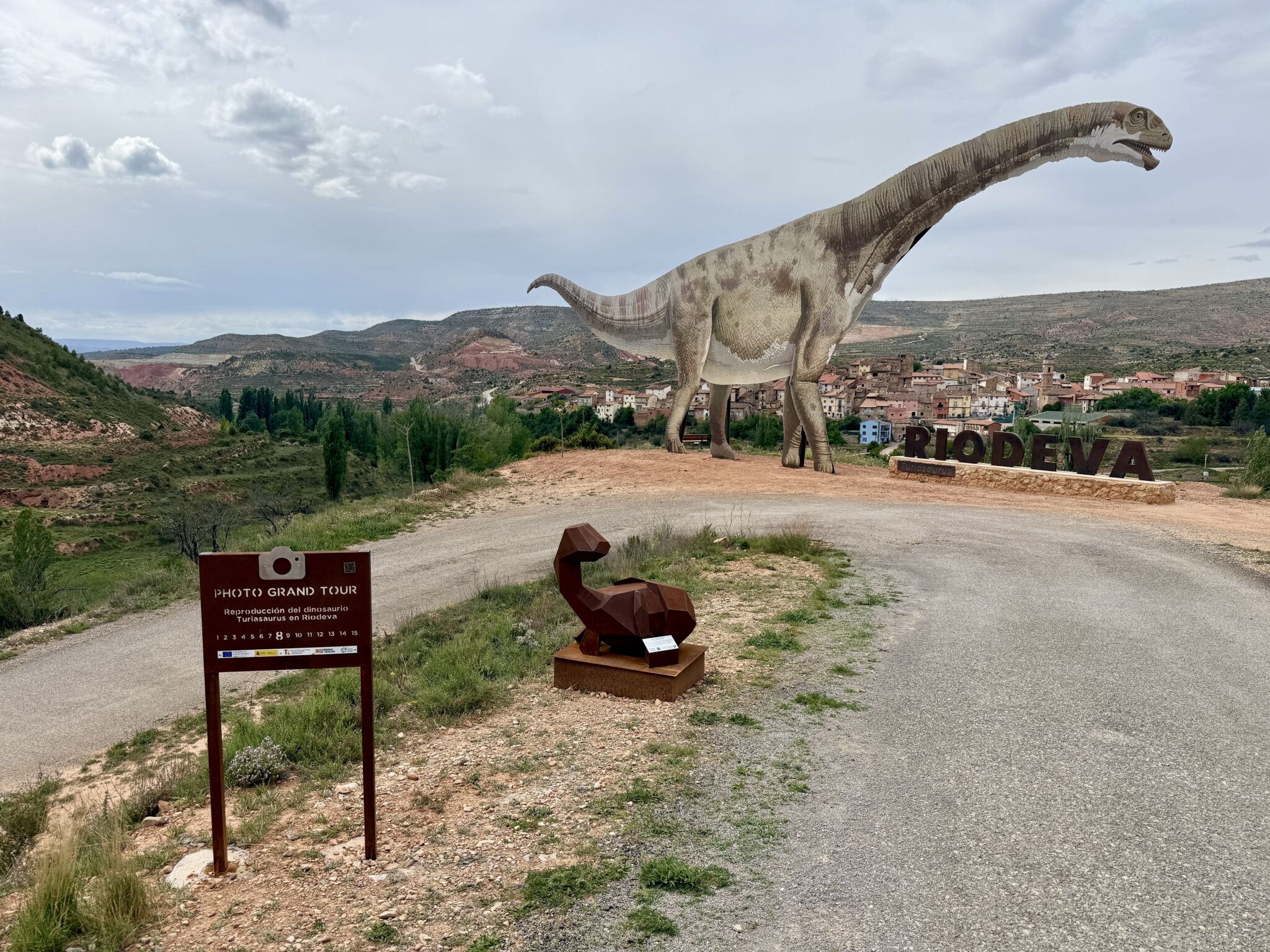 Una gran estatua de dinosaurio se alza cerca de la entrada de un pueblo llamado Riodeva, rodeado de colinas y vegetación que recuerda a Asturias. Una escultura geométrica de dinosaurio de color marrón y un cartel de recorrido fotográfico están junto a la carretera que lleva al pueblo.