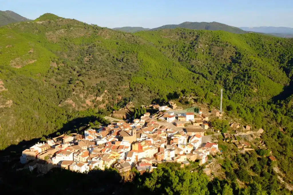 Un pequeño pueblo con tejados de tejas rojas está enclavado entre verdes colinas boscosas, rodeado de densos árboles y montañas bajo un cielo azul despejado.