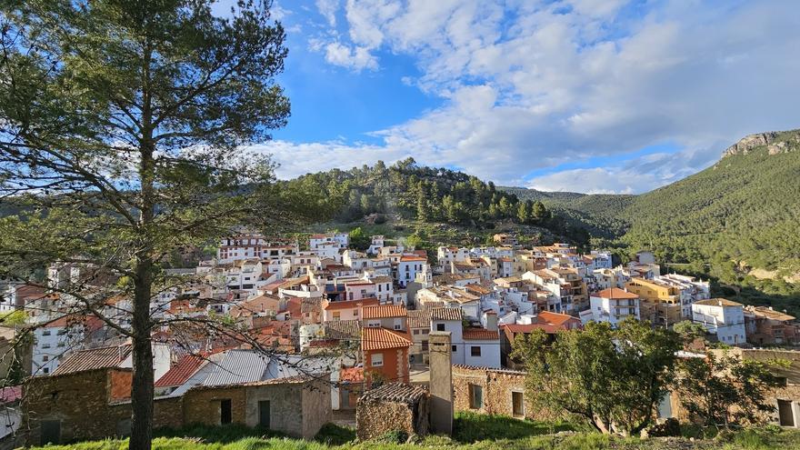 Al pie de una colina verde y arbolada, bajo un cielo azul parcialmente nublado y con árboles en primer plano, se alza un pintoresco pueblo con casas agrupadas de tejados blancos y de terracota.