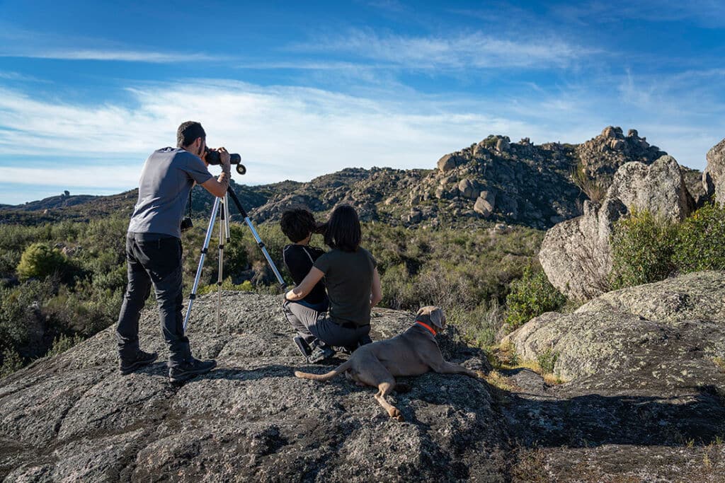 Un hombre utiliza unos prismáticos en un trípode mientras dos personas se sientan a su lado y un perro descansa cerca. Están en la cima de una colina rocosa que domina un paisaje montañoso bajo un cielo azul.