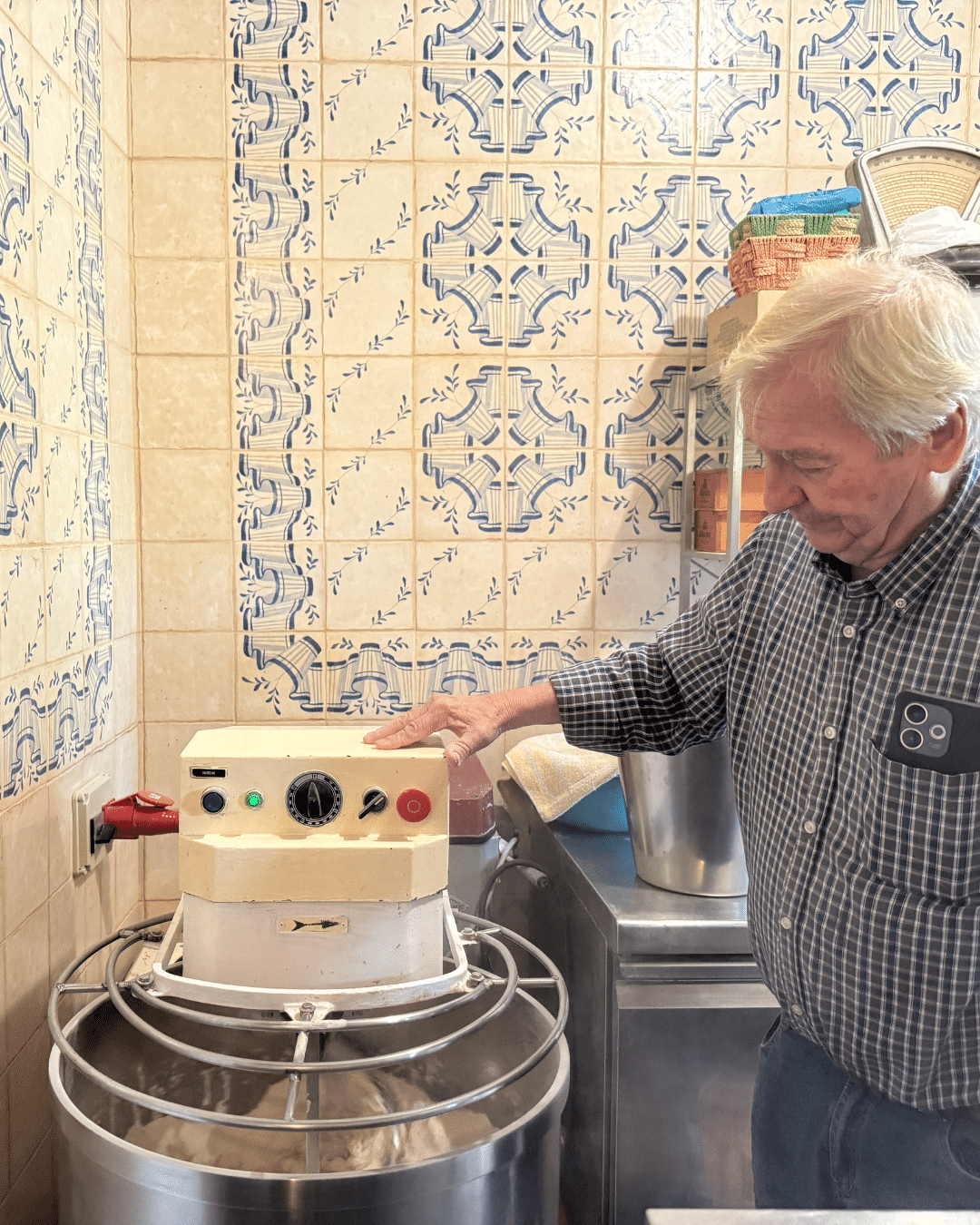 Un hombre mayor con el pelo blanco maneja una gran amasadora en una cocina de Asturias, rodeada de paredes de azulejos estampados y estanterías con productos de repostería.