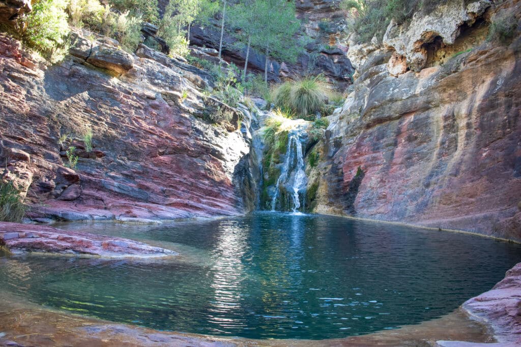 Una pequeña cascada cae en una piscina de roca transparente rodeada de escarpados acantilados de piedra multicolor y verde vegetación en un entorno natural tranquilo.