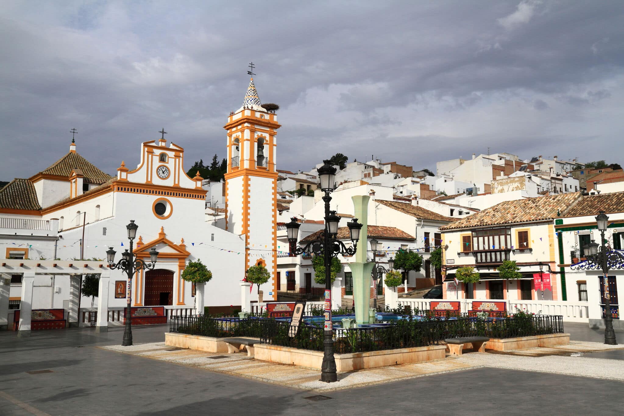 Una plaza de Asturias con una fuente central, bancos, farolas ornamentadas y árboles en macetas. Edificios blancos con tejados de tejas rojas rodean la plaza, incluida una iglesia con un campanario bajo un cielo nublado.