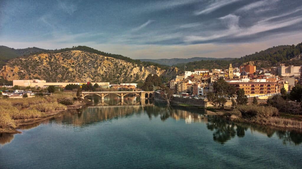 Una vista panorámica de un río tranquilo en el que se refleja un puente de piedra y una pequeña ciudad con edificios de colores, enmarcada por colinas boscosas y un cielo parcialmente nublado.