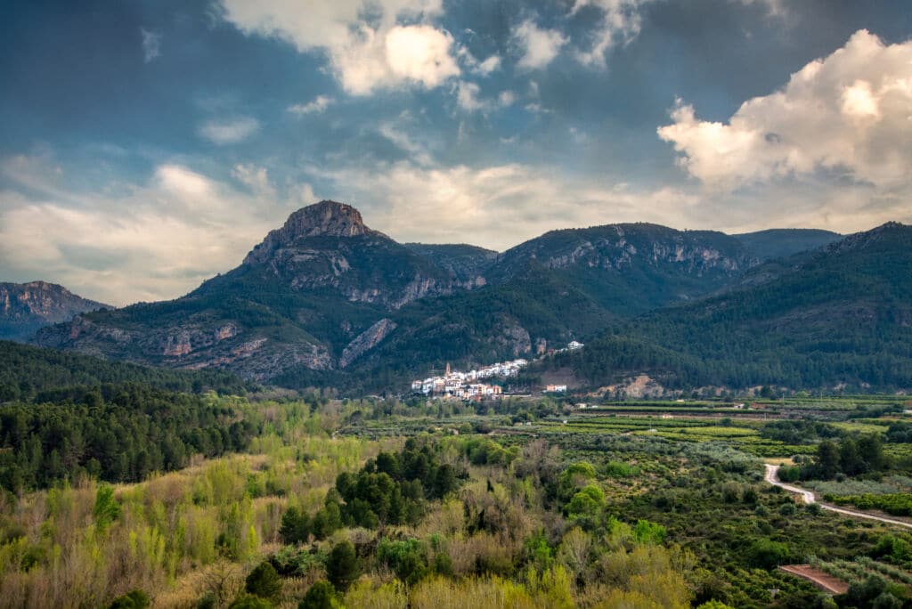 Un exuberante valle verde con árboles y arbustos dispersos, que conduce a un pequeño pueblo enclavado en la base de altas y escarpadas montañas bajo un cielo parcialmente nublado.