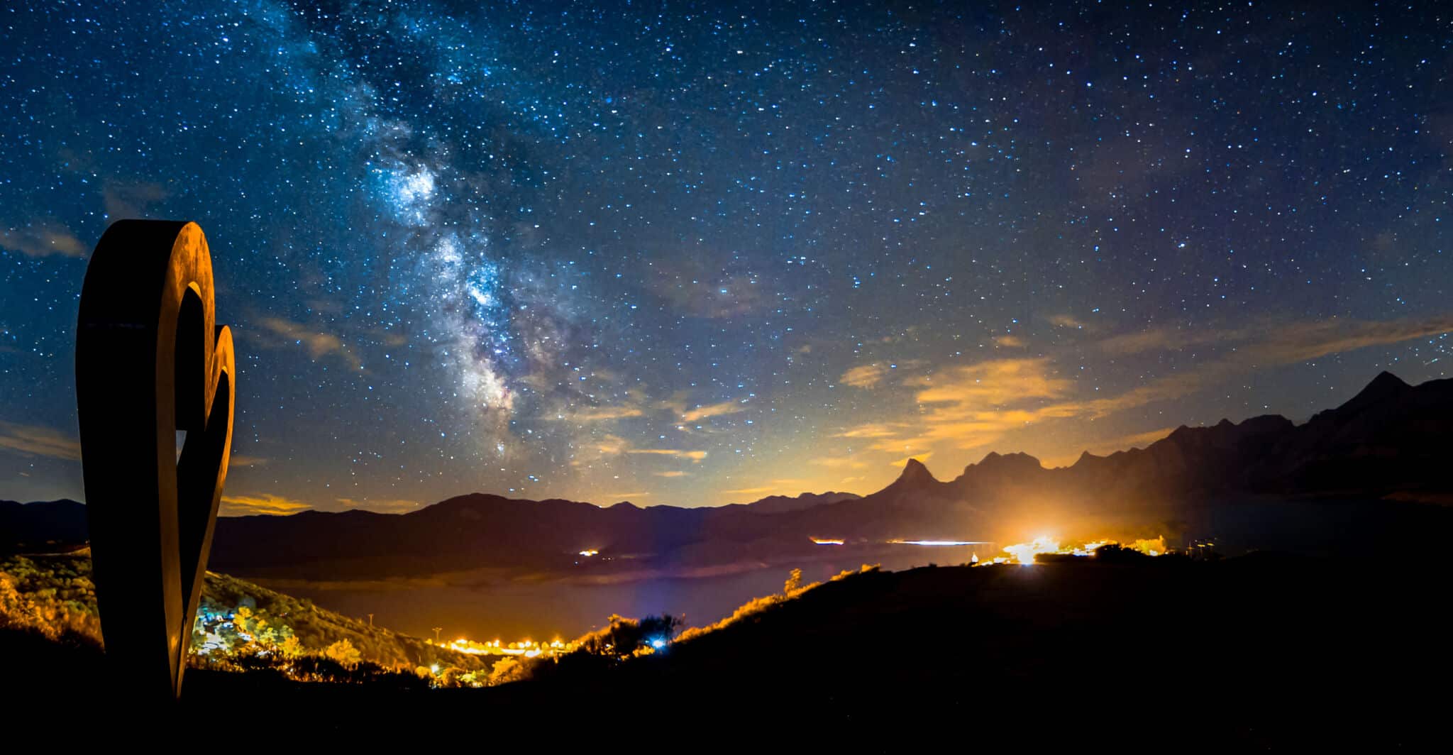 Un cielo nocturno lleno de estrellas con la Vía Láctea sobre montañas lejanas y un lago, con una ciudad resplandeciente debajo y una gran escultura de un corazón silueteada en primer plano.