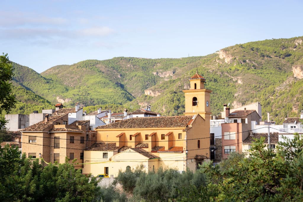 Una iglesia amarilla con un campanario se alza entre las casas, con un telón de fondo de verdes colinas boscosas bajo un cielo despejado.
