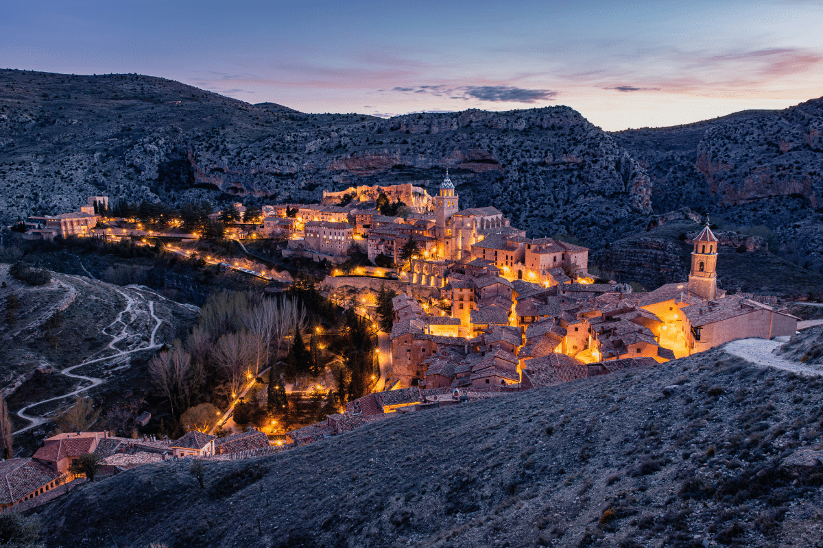 Un pueblo en lo alto de una colina brilla con luces cálidas al atardecer, rodeado de colinas escarpadas y una carretera serpenteante. Edificios de piedra, calles estrechas y la torre de una iglesia son visibles bajo un cielo azul y rosa que se desvanece.