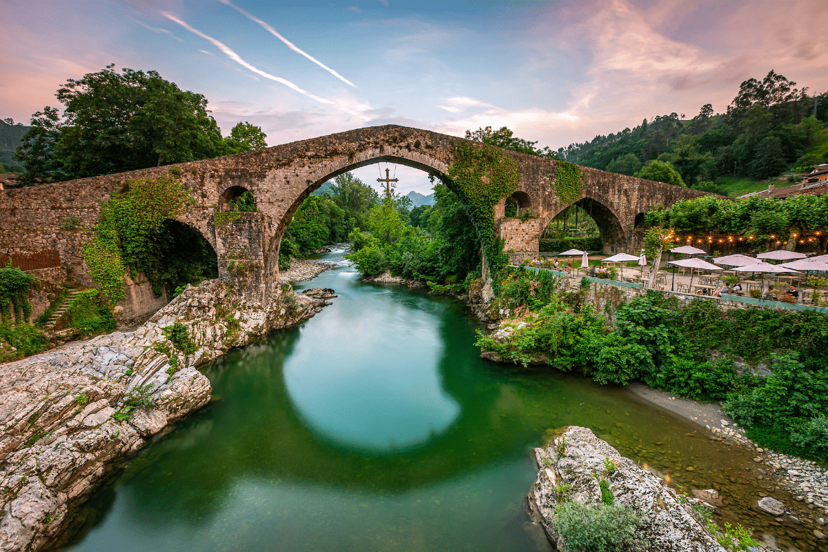 Un antiguo puente de piedra con grandes arcos atraviesa un río tranquilo y verde rodeado de frondosos árboles y orillas rocosas al atardecer; una cruz cuelga del puente, y a la derecha se ve una terraza ribereña con sombrillas.