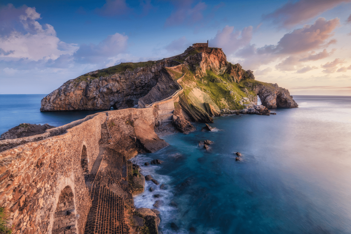 Un sinuoso camino de piedra conduce a una pequeña capilla en lo alto de una isla rocosa rodeada de un mar azul en calma, bajo un cielo colorido al atardecer con nubes dispersas.