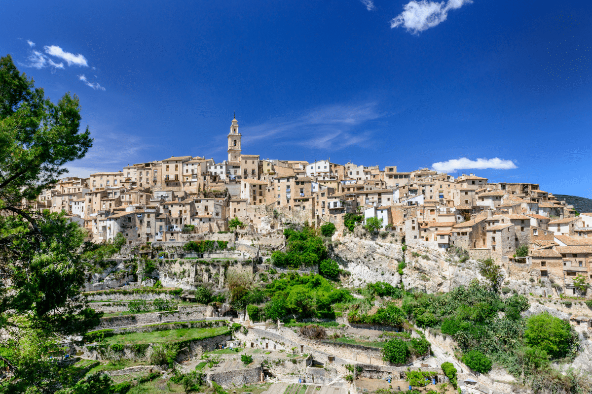 Un pueblo en lo alto de una colina con edificios de piedra y una prominente torre de iglesia domina los verdes jardines en terrazas bajo un cielo azul brillante con nubes dispersas. Árboles y terreno rocoso rodean el pintoresco pueblo.