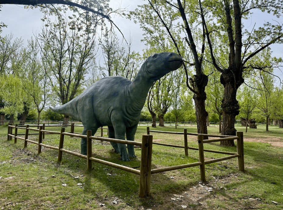 Una gran estatua de dinosaurio verde se encuentra dentro de una zona vallada de madera en un parque con césped verde y árboles altos y frondosos en Asturias en un día nublado.