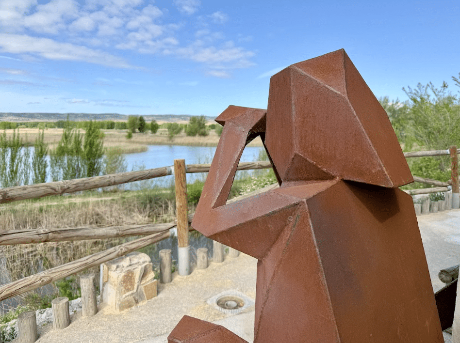 Una escultura geométrica metálica de un mono, de color óxido, se asienta junto a una valla de madera en Asturias, con vistas a un estanque con árboles y hierba bajo un cielo azul parcialmente nublado.