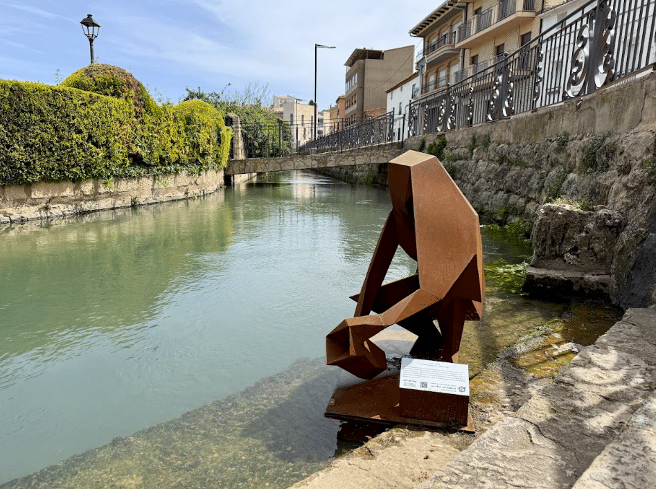Una escultura metálica oxidada y angulosa de una figura sentada descansa junto a un canal de aguas tranquilas y muros de piedra en Asturias, rodeada de vegetación. Al fondo, edificios residenciales y barandillas de hierro bajo un cielo azul despejado.
