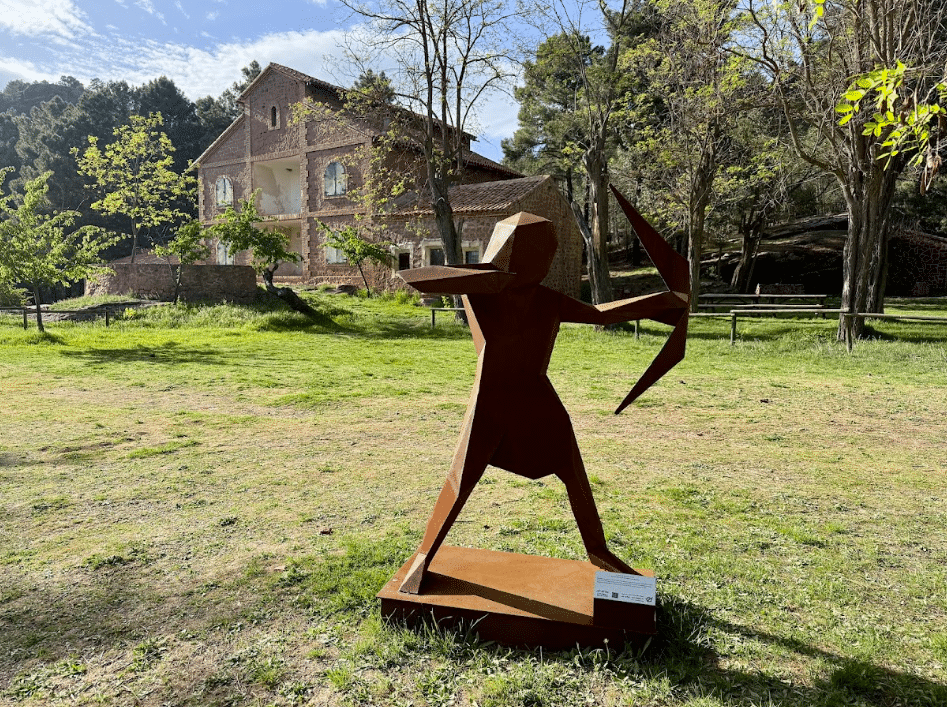 Una escultura geométrica de metal, de color óxido, que representa a un arquero con un arco, se alza sobre la hierba de un parque de Asturias, con un edificio de ladrillo y árboles al fondo, bajo un cielo parcialmente nublado.