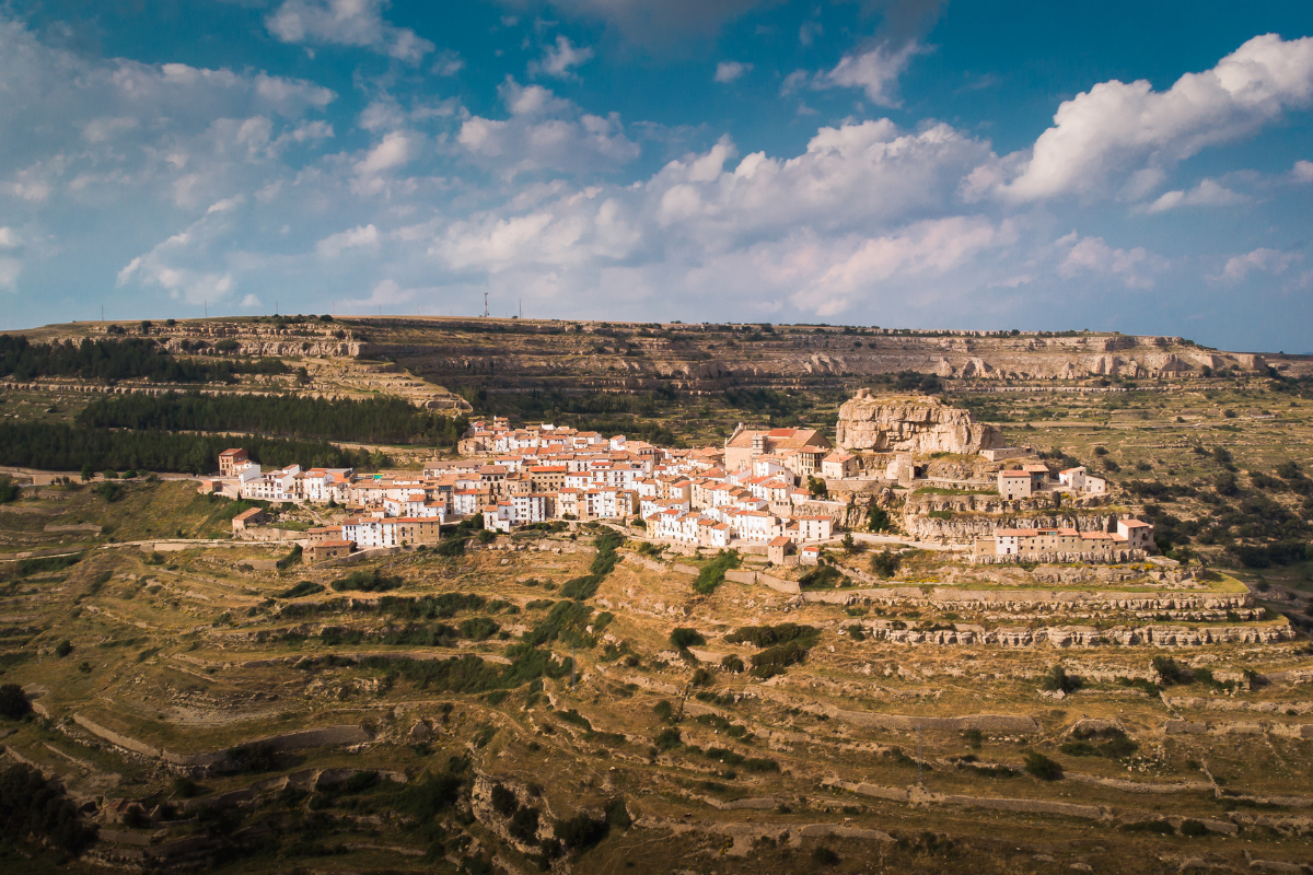 Vista aérea de un pueblo en la ladera de una colina, con edificios blancos y rojos agrupados, rodeado de terreno rocoso en terrazas y manchas verdes bajo un cielo azul parcialmente nublado.