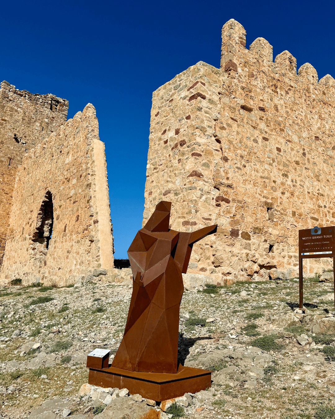Una escultura metálica geométrica de una mano, de color óxido, señala hacia el exterior frente a los muros de un antiguo castillo de piedra bajo un cielo azul despejado en Asturias, con una señal marrón cerca sobre un terreno rocoso.