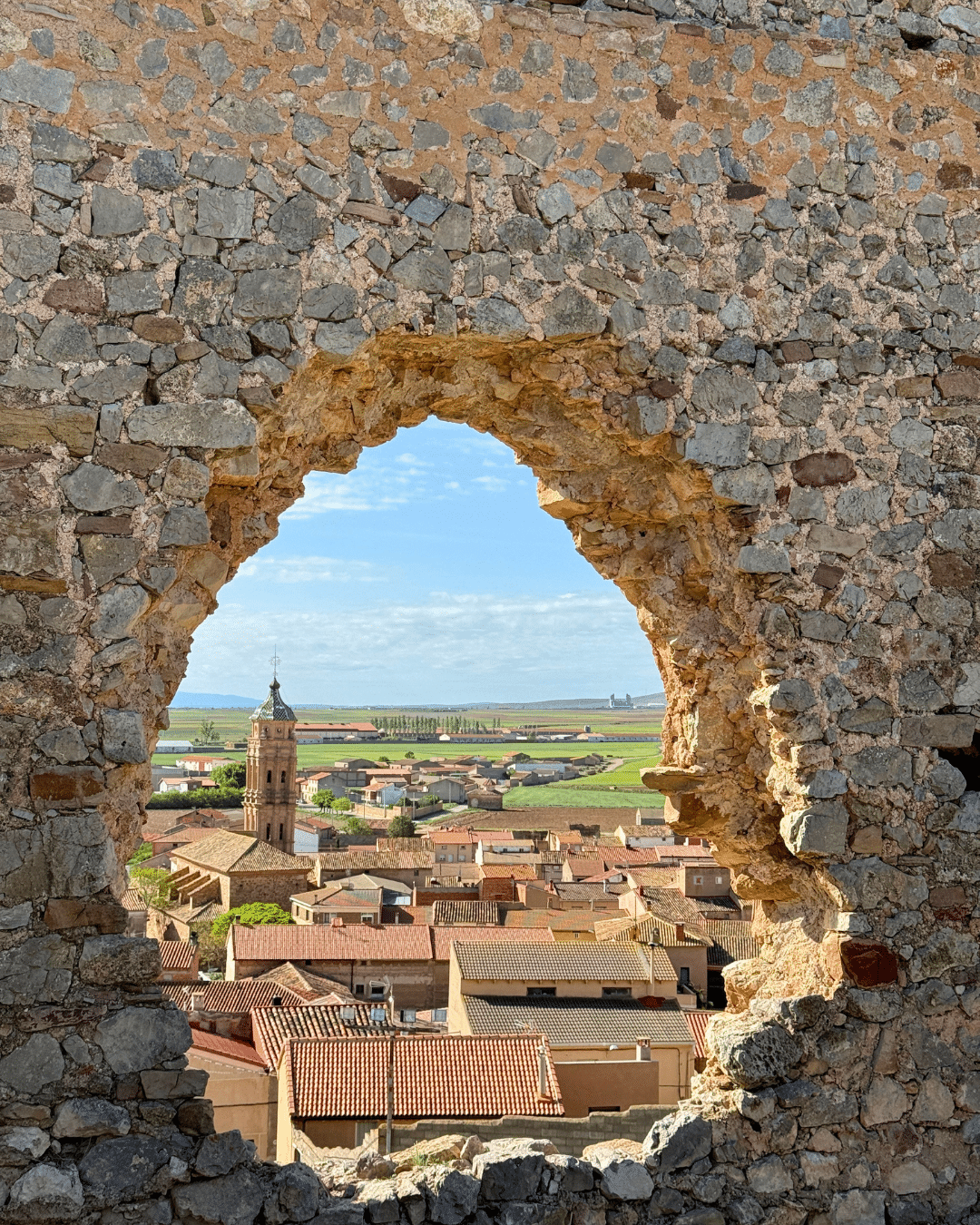 Un muro de piedra con una gran abertura en arco enmarca un encantador pueblo asturiano, con tejados de tejas rojas, una torre de iglesia, campos verdes y un cielo azul de fondo.